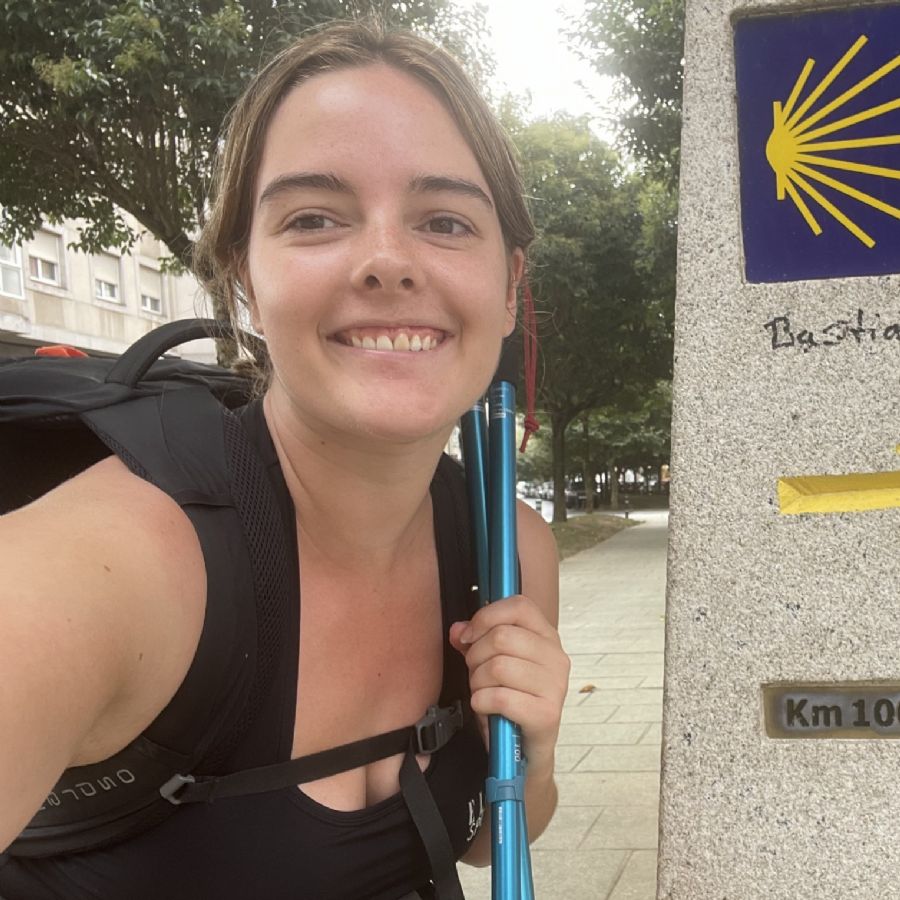 Woman with a backpack and hiking poles standing next to a Camino de Santiago marker.