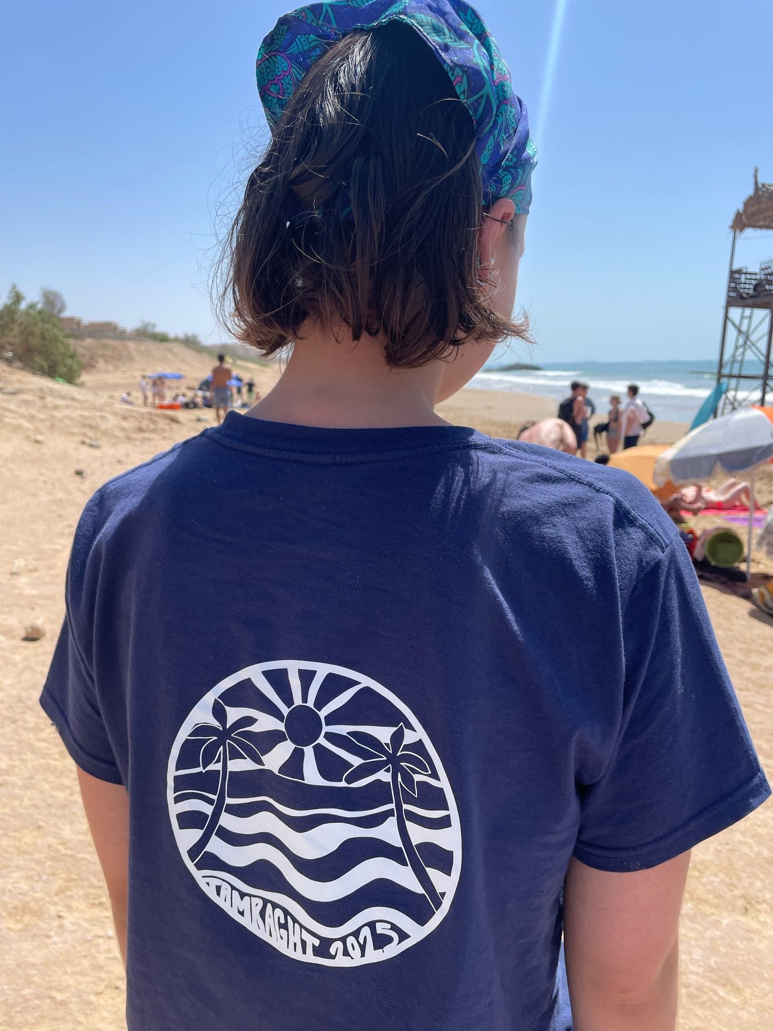 Person wearing a blue t-shirt with a beach-themed design on a sandy beach.