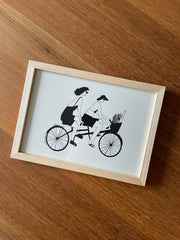 Framed artwork of two people riding a tandem bicycle.