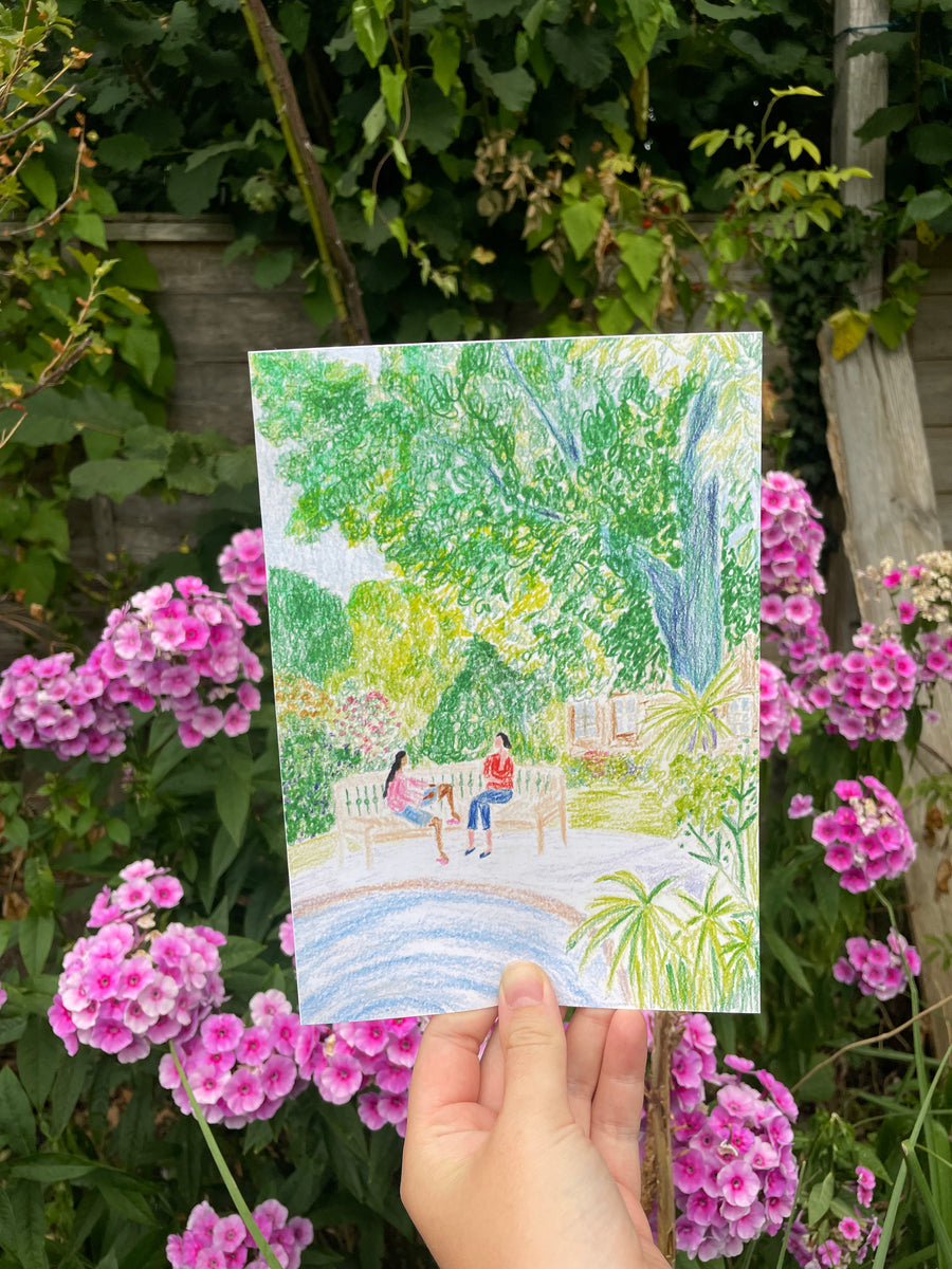 A hand holds a square colored pencil drawing of a park scene with trees, benches, and two people sitting by a pond, set against a real garden background with pink flowers and green foliage.