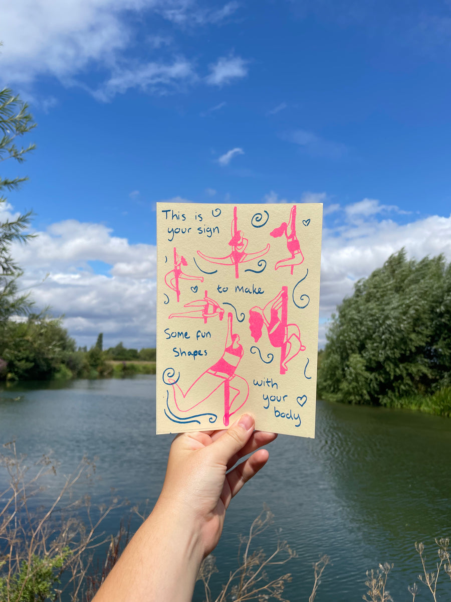 hand holding a riso print with pink and blue hand-drawn figures and handwritten text in front of a lake and trees under a blue sky.