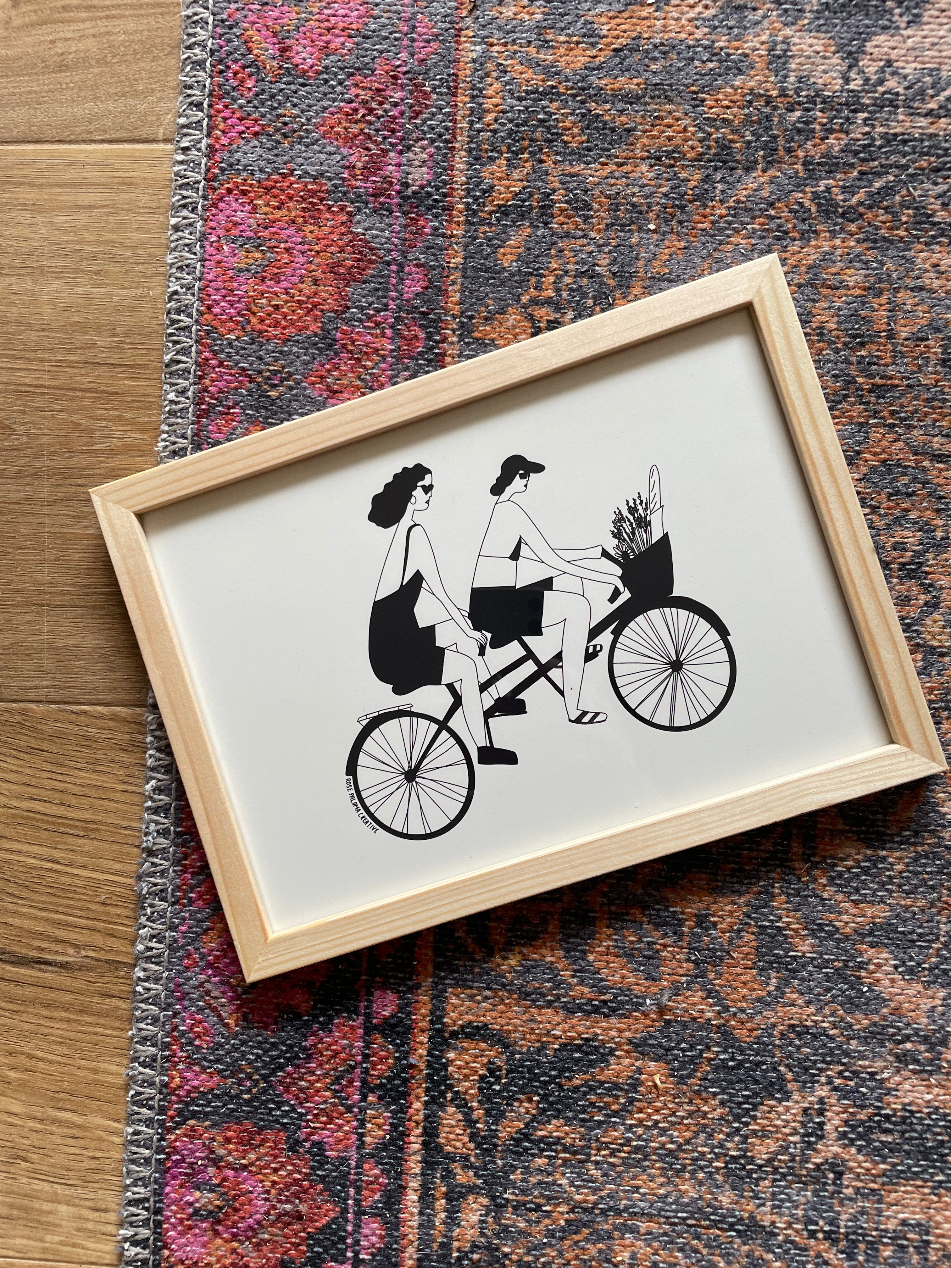 Framed artwork of two people on a tandem bicycle.