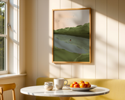 Framed landscape painting on a wall above a table with a cup and plate of oranges.